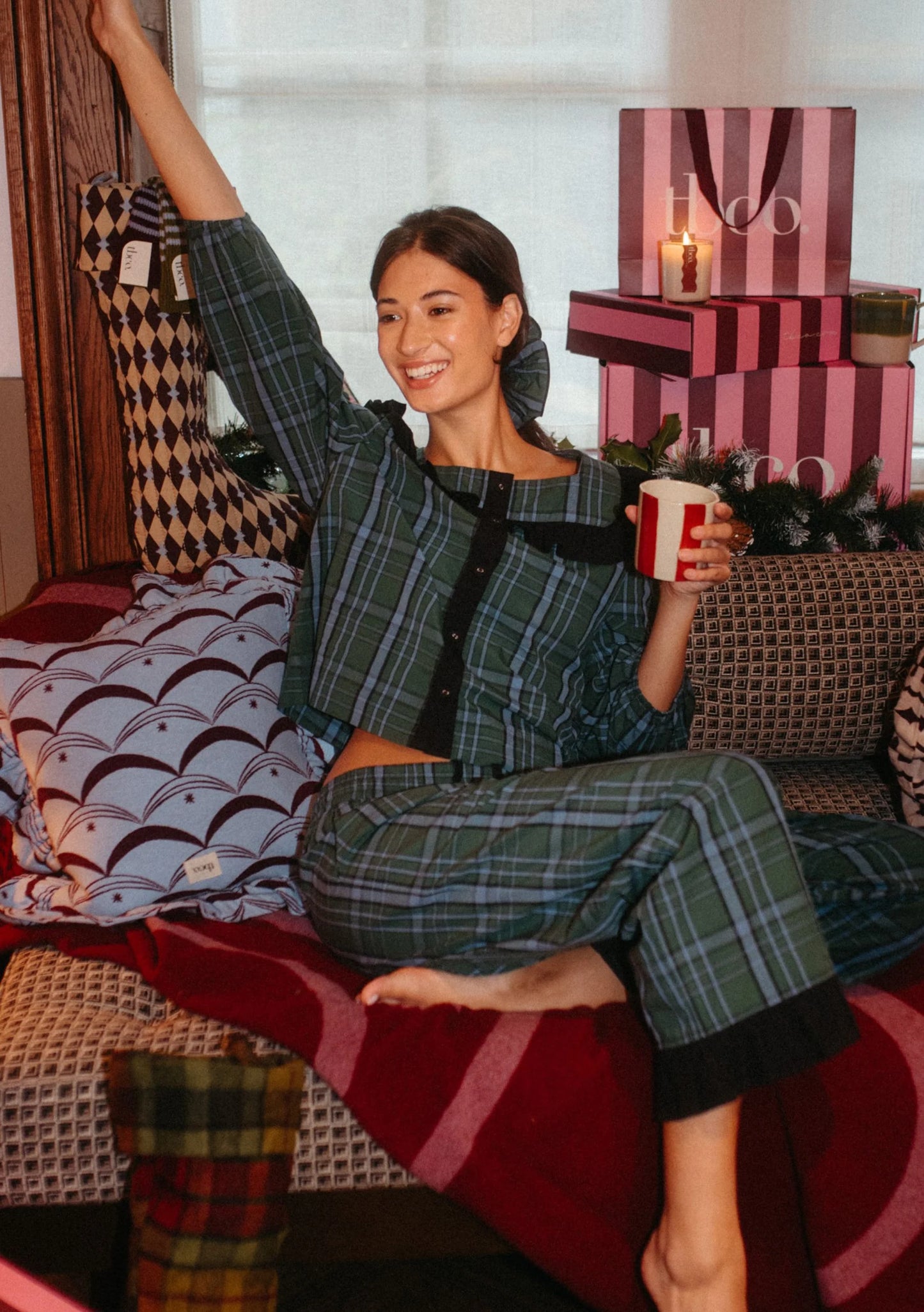 Woman in plaid pajama set holding a red mug in a cozy room with gift boxes. Pyjamas are by TBCo and are filtered by Shop My Palette for the Deep Winter, Cool Winter, and Clear Winter colour seasons.
