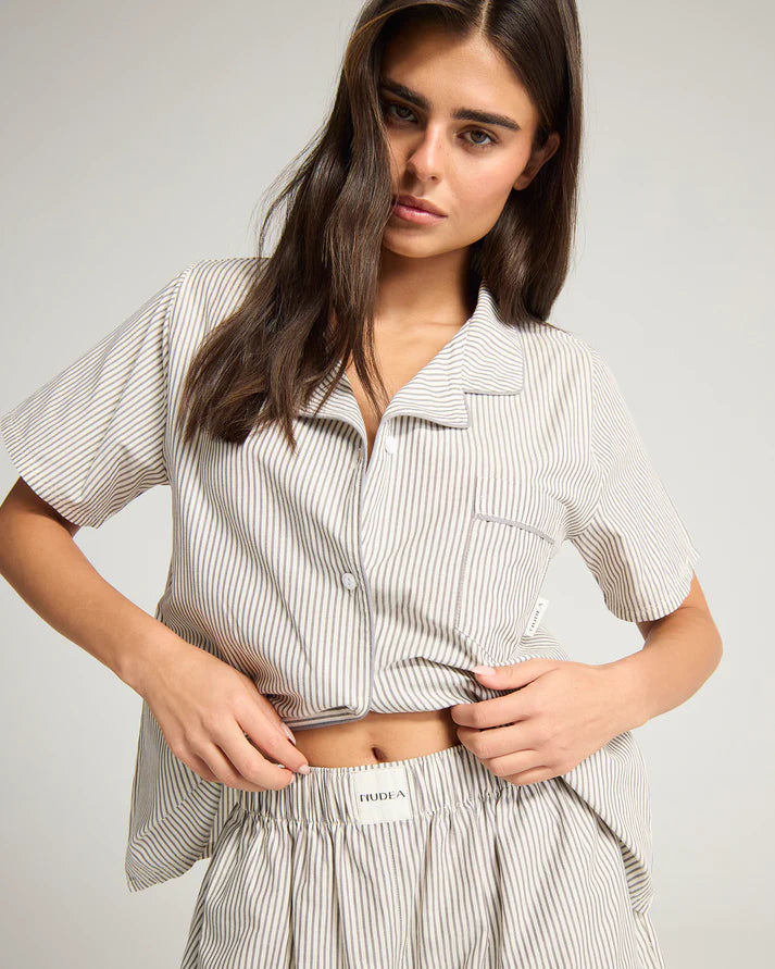 Model wearing short sleeved pyjama top and shorts, in grey and white striped BCI cotton with piped details. Pyjamas are by Nudea and are filtered for the Cool Summer, Soft Summer, and Light Summer colour seasons by Shop My Palette.