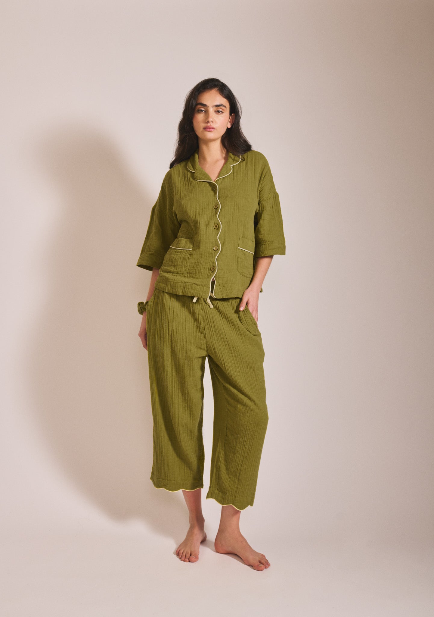 Model wearing a relaxed moss green pyjama set with short sleeves, button front, contrast piping detail, and loose cropped trousers.