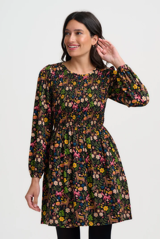 A woman modeling a long-sleeve smocked dress with a busy multicolor floral and deer print on a black background, set against a plain light gray studio backdrop.