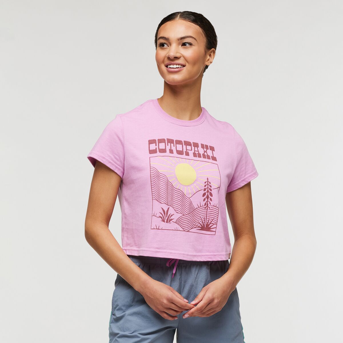 Bright pink cropped T-shirt with Cotopaxi and a sunrise logo, made from organic cotton-recycled polyester fabric blend.