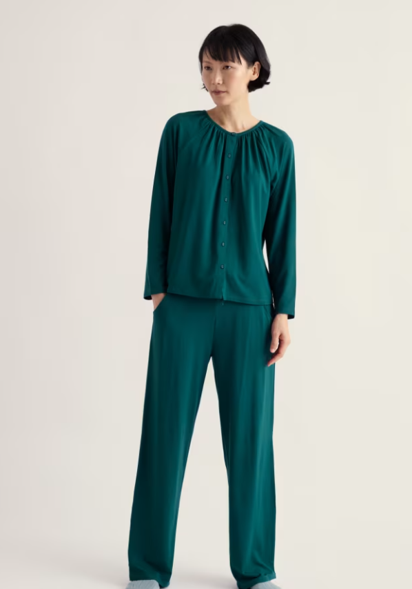 A woman modelling a matching two-piece loungewear set in deep teal green, featuring a long-sleeve button-front top and wide-leg pants, against a plain off-white studio background.
