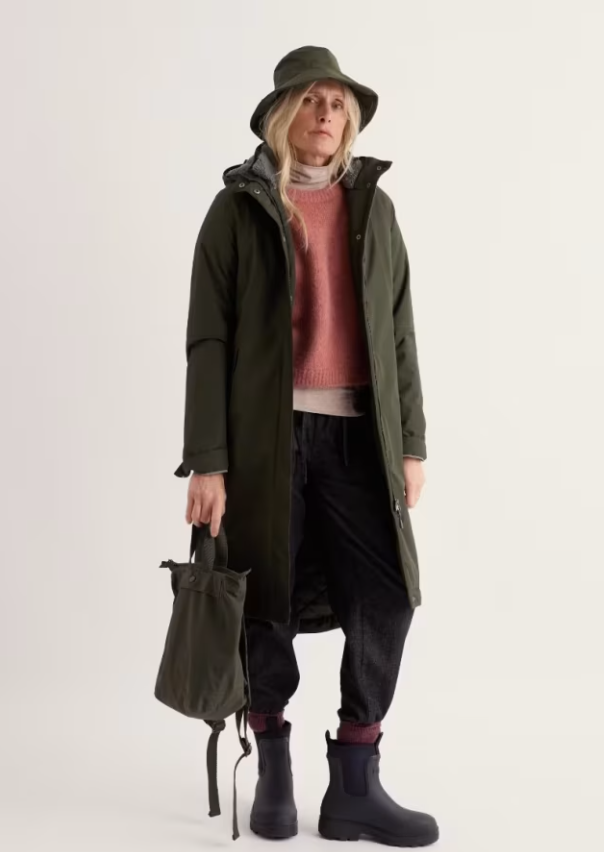 Model wearing a deep olive green raincoat and hat over a coral sweater and dark trousers.