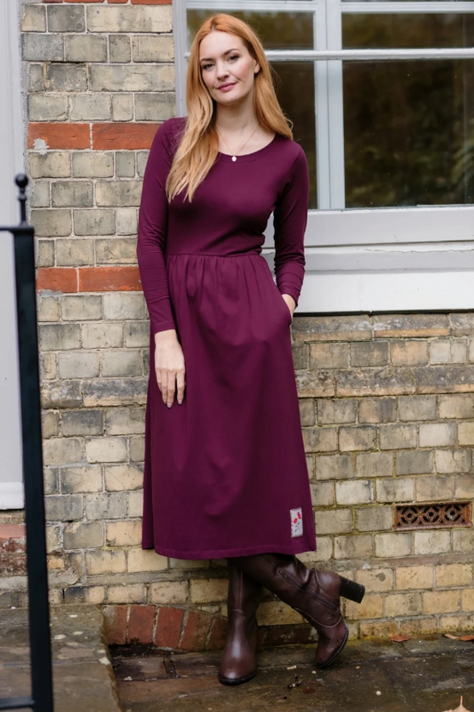 A woman wearing a long-sleeve midi dress in deep burgundy with brown leather boots, standing against a brick wall next to a window.