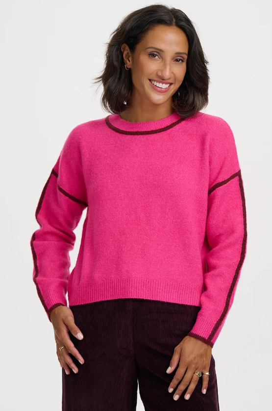 A woman wearing a bright pink knit sweater with dark burgundy contrast trim along the crew neck, cuffs, and sleeves, paired with dark burgundy pants, against a plain white studio background.