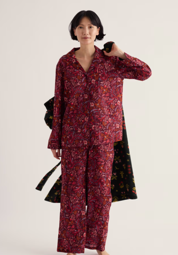 A woman modelling a two-piece pyjama set with a red and black floral paisley pattern, holding a black robe with a matching floral print against a plain light background.