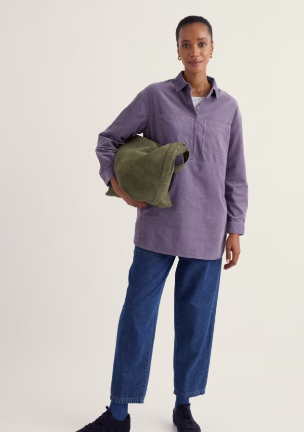 A model stands against a plain light background wearing a lavender-purple long-sleeve button-up shirt, blue jeans, and black shoes, while holding an olive green bucket bag.
