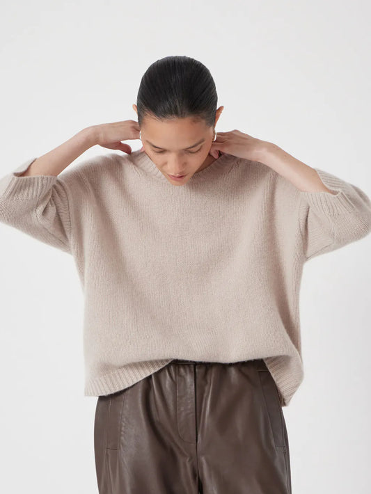 Soft taupe-beige relaxed cashmere knit sweater with a round neckline and elbow-length sleeves, styled with brown leather trousers.