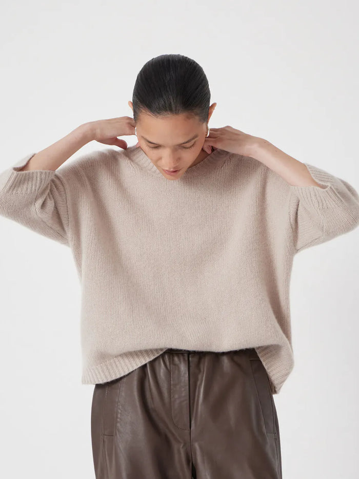 Soft taupe-beige relaxed cashmere knit sweater with a round neckline and elbow-length sleeves, styled with brown leather trousers.