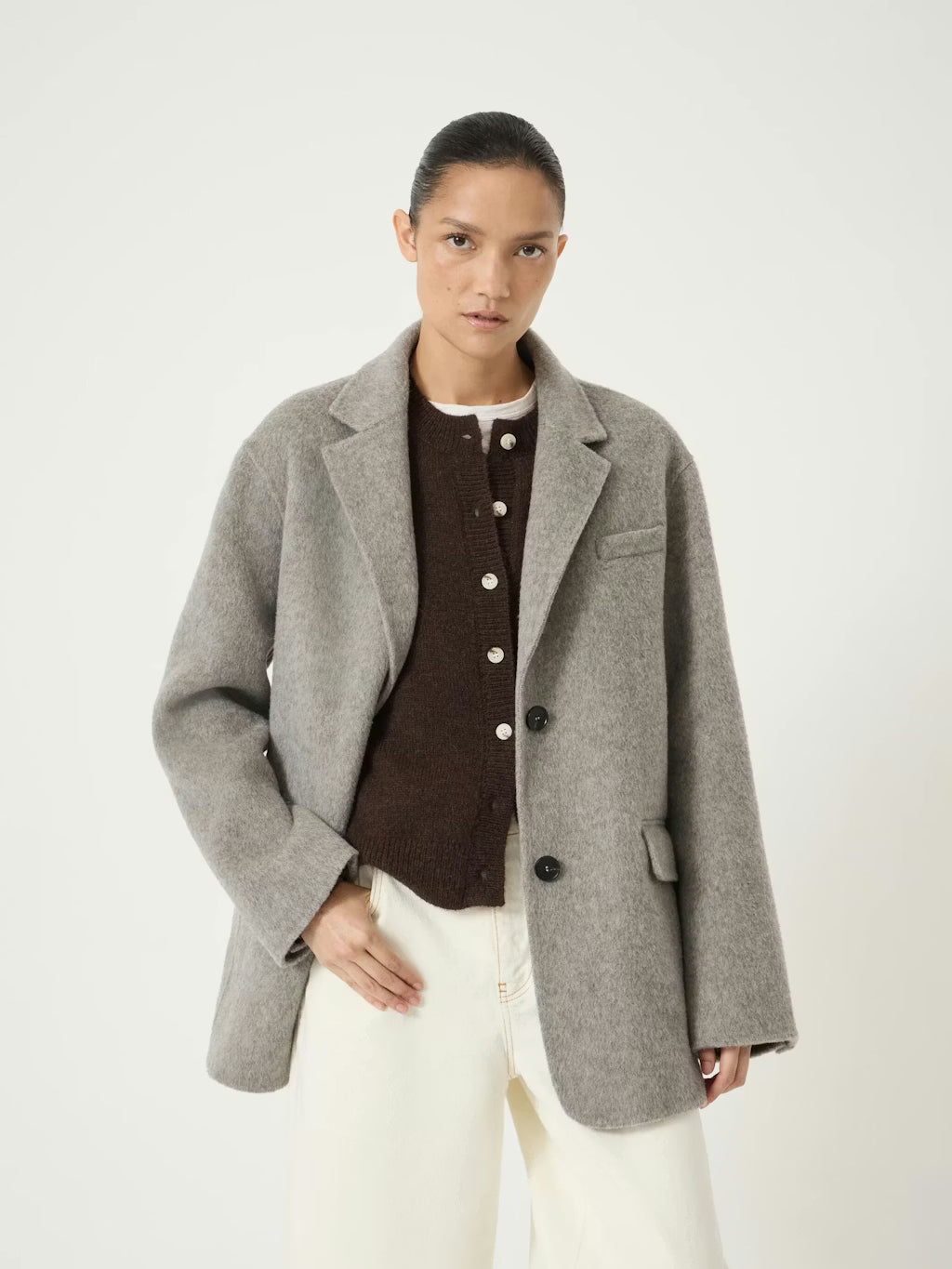 A model wearing a light gray wool-blend blazer over a dark brown buttoned cardigan and cream trousers, photographed against a clean off-white studio background.