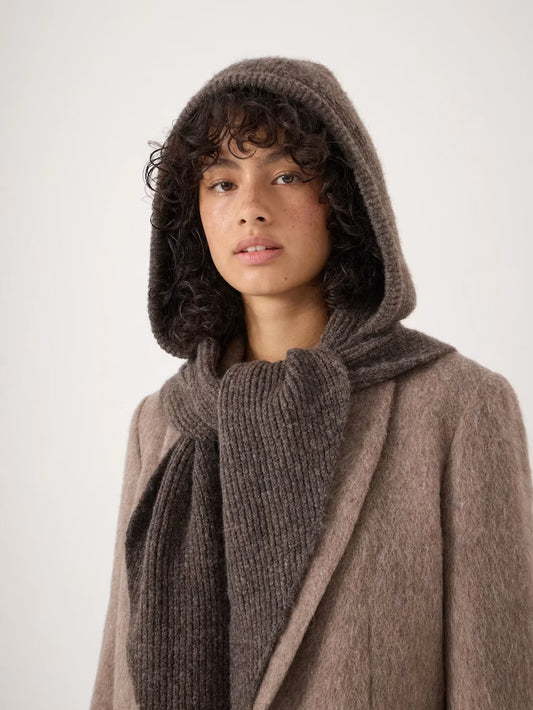 A model wearing a cozy brown knit hooded knit scarf, and a soft taupe-brown coat against a plain light background.