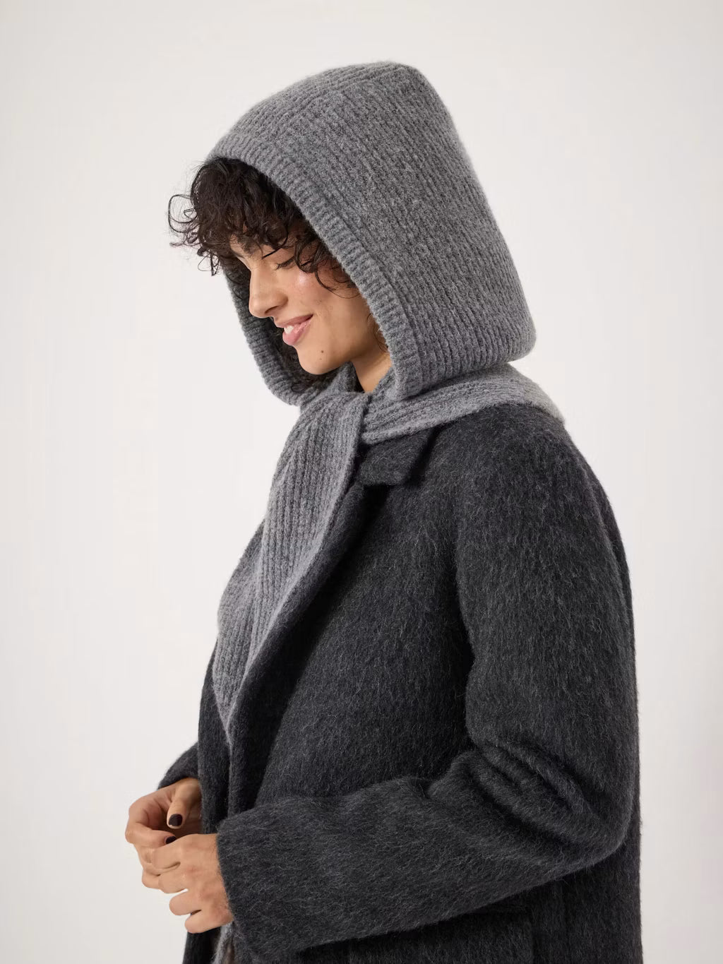 A woman wearing a dark charcoal gray textured coat with a wide ribbed knit hood and matching scarf, shown in profile against a plain light background.