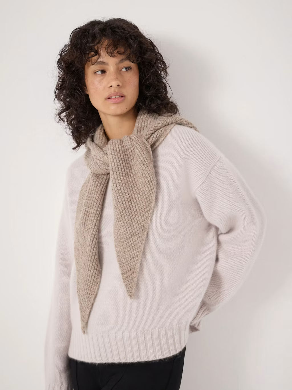 A model with curly dark hair is wearing a light pink knit sweater and a chunky beige scarf, photographed against a plain off-white wall.