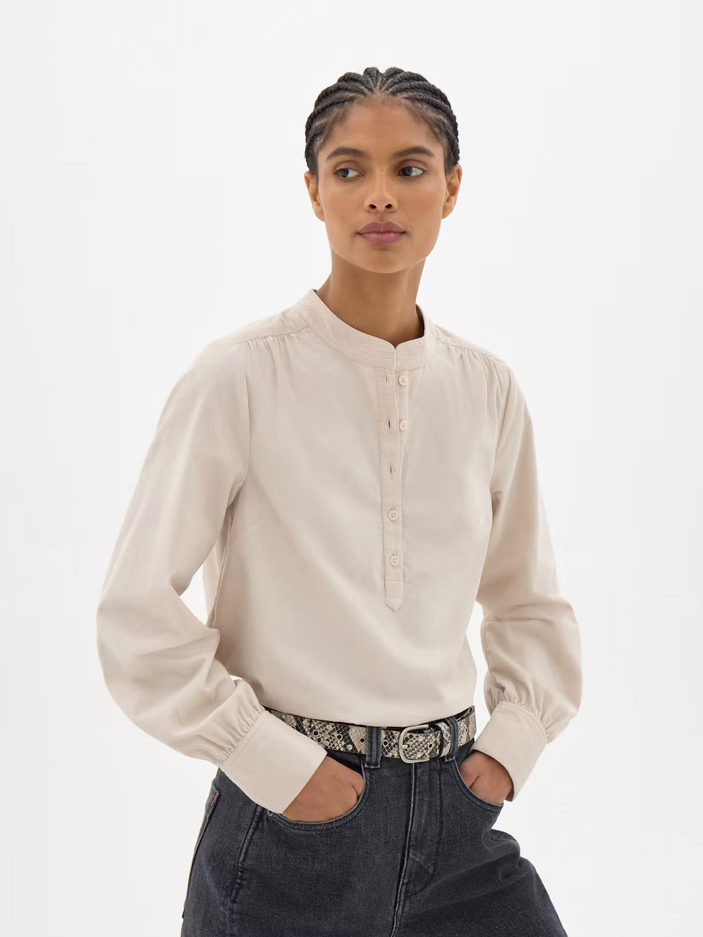 A woman modeling a light beige long-sleeve blouse with a buttoned mandarin collar and gathered cuffs, paired with dark jeans and a patterned belt, against a plain white studio background.