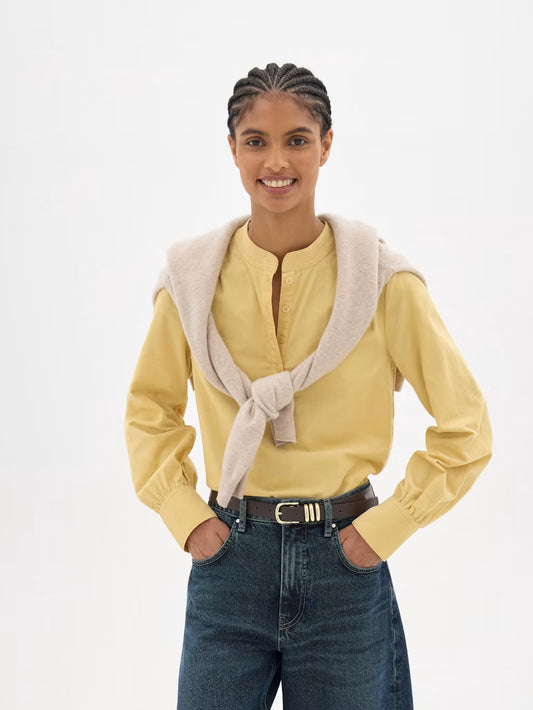 A woman modeling a pale yellow long-sleeve blouse with gathered cuffs, paired with a light beige knit sweater draped over her shoulders and blue jeans, against a plain white studio background.