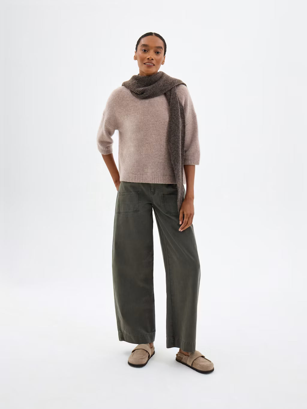 A model stands against a plain white studio background wearing a light taupe knit sweater, dark olive wide-leg trousers, a dark brown scarf, and taupe flat sandals.