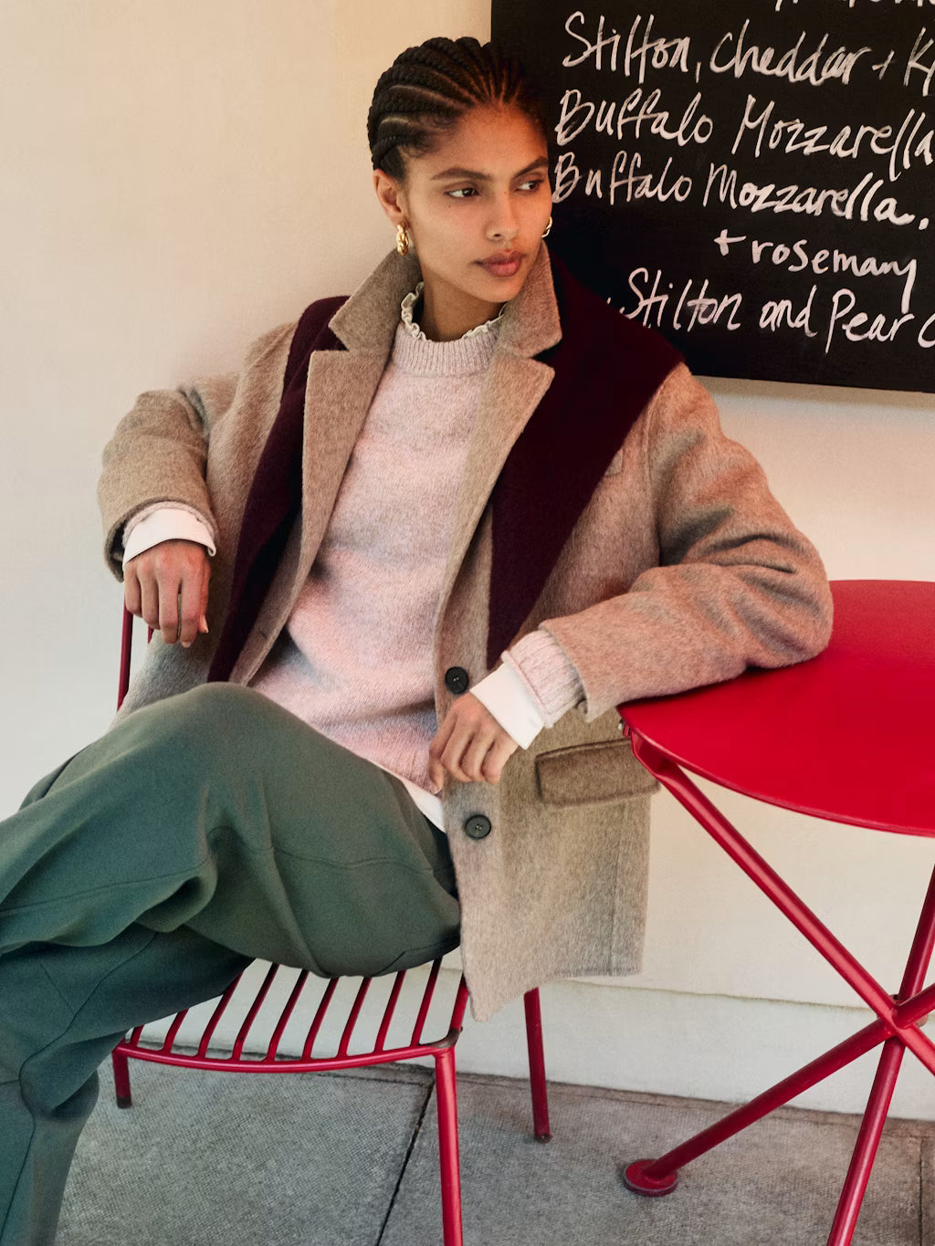 A model is seated on a red metal chair in an indoor setting, wearing a beige textured coat with dark burgundy lapels over a light pink knit sweater and olive green trousers. A red table is next to her, and a chalkboard menu is visible in the background.