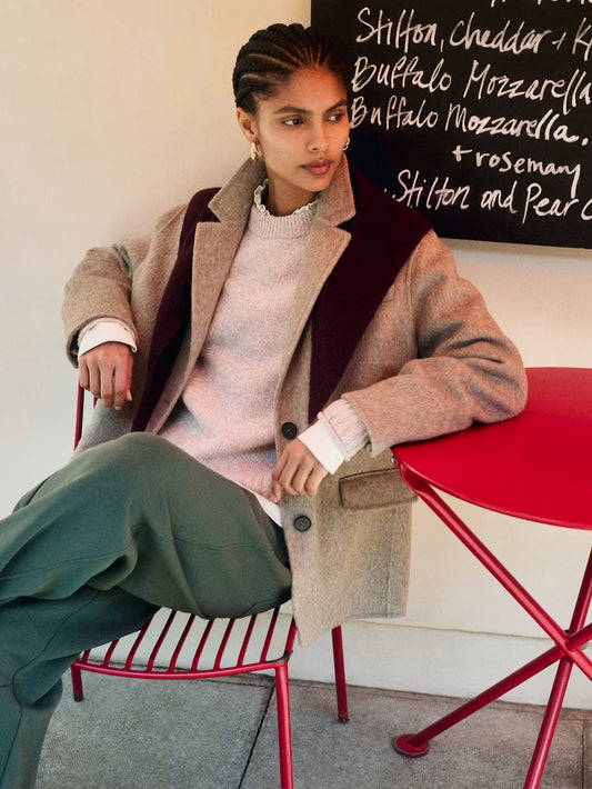 A model is seated on a red metal chair in an indoor setting, wearing a beige textured coat with dark burgundy lapels over a light pink knit sweater and olive green trousers. A red table is next to her, and a chalkboard menu is visible in the background.