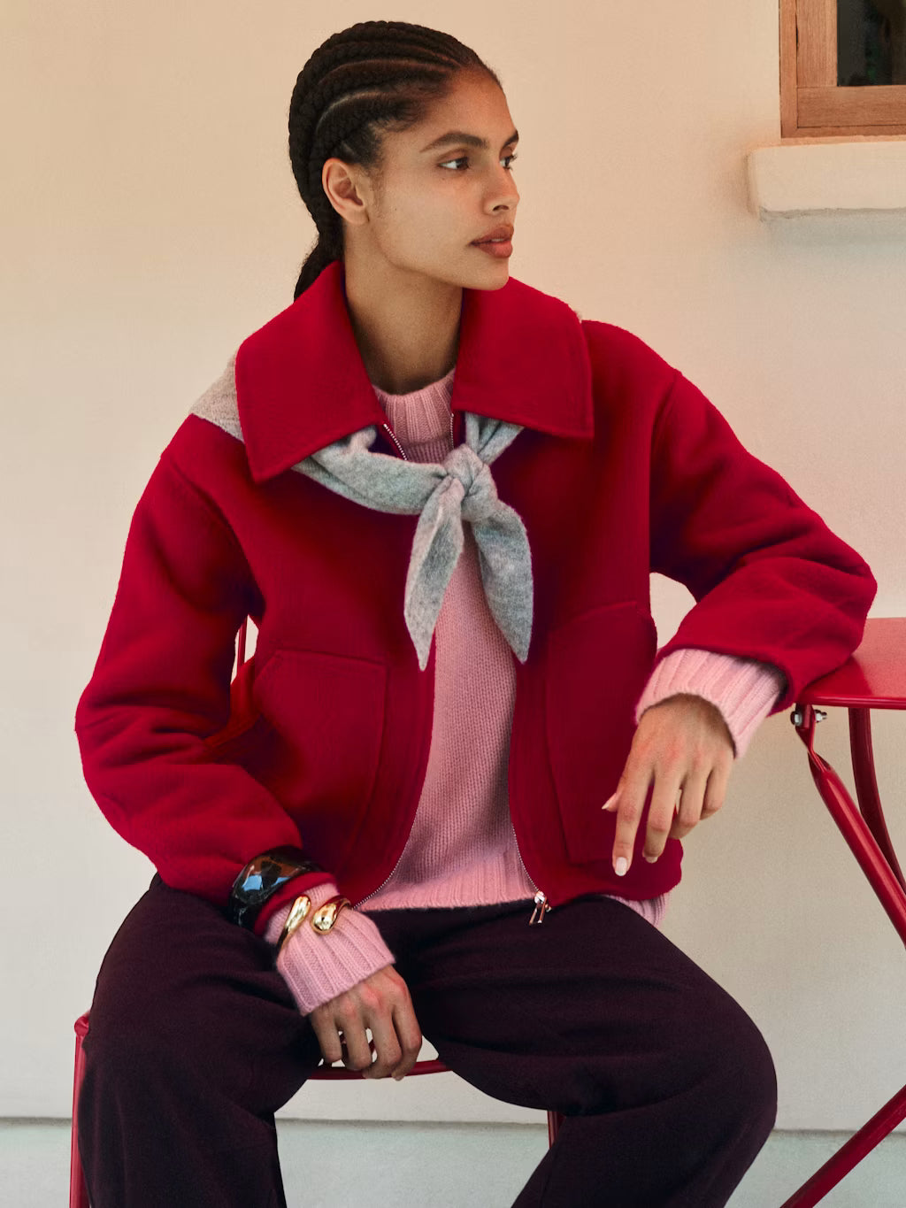 A model wearing a vibrant red cropped jacket with a high collar, layered over a pink sweater and a light gray necktie, paired with dark trousers, sitting on a red chair.