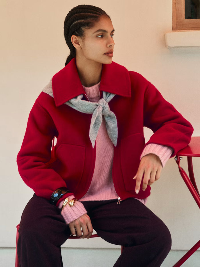 A model wearing a vibrant red cropped jacket with a high collar, layered over a pink sweater and a light gray necktie, paired with dark trousers, sitting on a red chair.