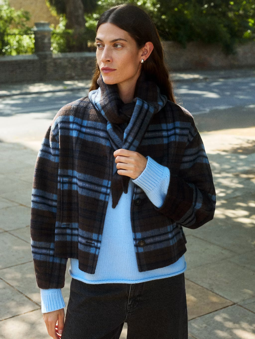 A woman wearing a dark brown and blue plaid wrap jacket over a light blue ribbed sweater, standing outdoors on a paved walkway.