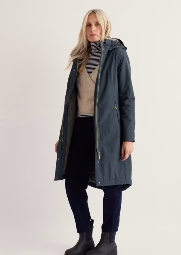 A woman modeling a long hooded parka coat in a muted blue-gray color, worn open over a striped turtleneck, beige sweater, dark pants, and black boots, against a plain light studio background.