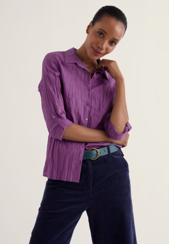A woman modeling a purple crinkled cotton button-up shirt with three-quarter sleeves, paired with dark navy corduroy trousers and a teal belt, against a plain off-white studio background.