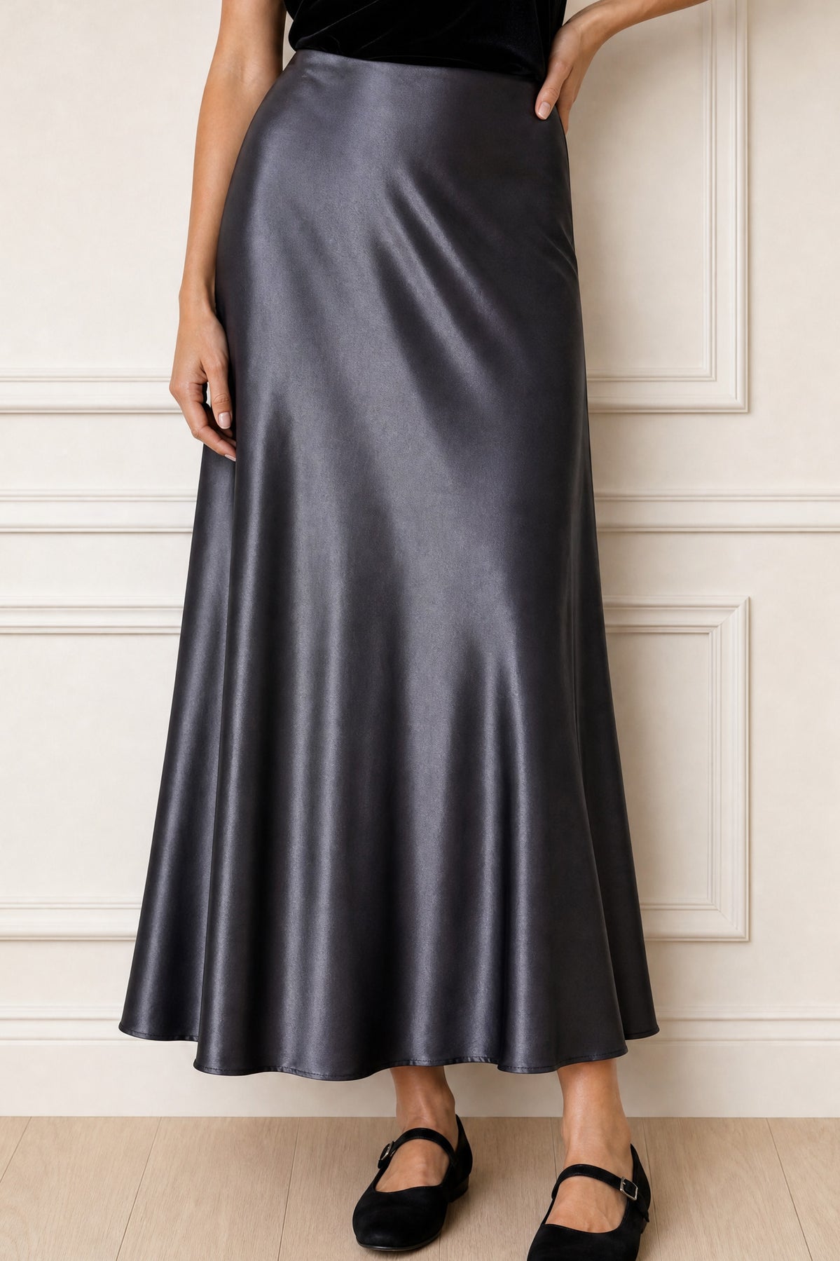A woman wearing a high-waisted, floor-length satin midi skirt in a dark gray color, paired with black flats, standing against a white paneled wall.