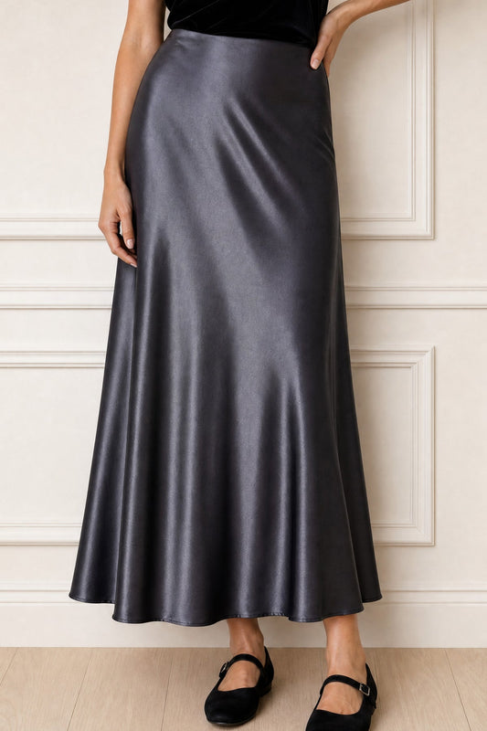 A woman wearing a high-waisted, floor-length satin midi skirt in a dark gray color, paired with black flats, standing against a white paneled wall.