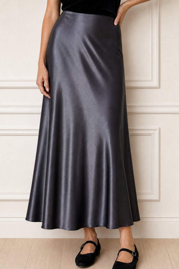 A woman wearing a high-waisted, floor-length satin midi skirt in a dark gray color, paired with black flats, standing against a white paneled wall.