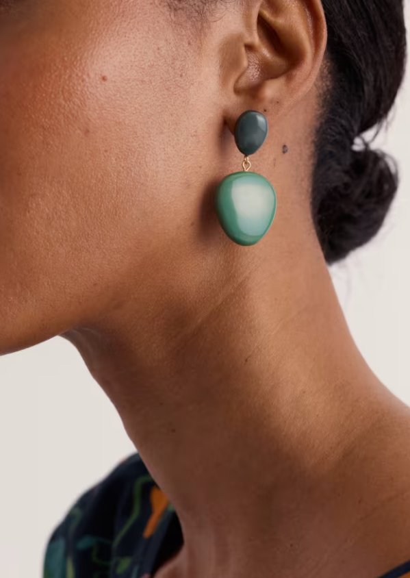 A close-up side profile of a model wearing a two-tone drop earring. The earring features a smaller dark green stone at the top and a larger, smooth matte green stone below, suspended by a small gold-tone ring.