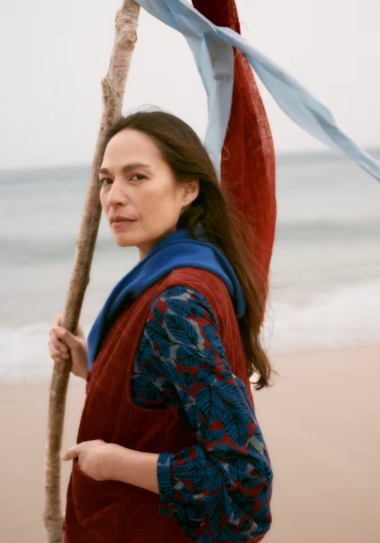 Woman on a beach wears rust colour gilet over a blue dress, with blue scarf.