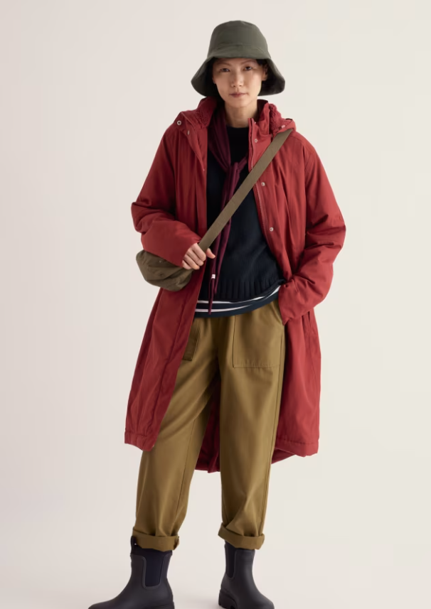 A model stands against a plain light background wearing a long red hooded coat over a dark top, olive pants, a bucket hat, and dark boots, while carrying a tan shoulder bag.