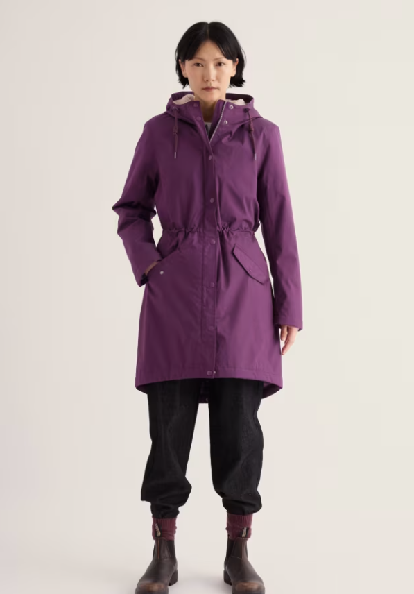 A model stands against a light off-white studio background wearing a mid-length purple hooded raincoat with a drawstring waist, black pants, and dark brown boots.