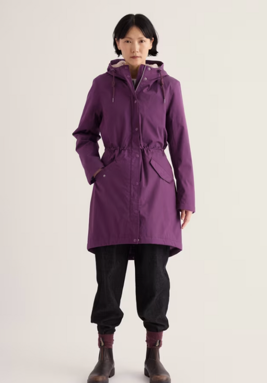 A model stands against a light off-white studio background wearing a mid-length purple hooded raincoat with a drawstring waist, black pants, and dark brown boots.