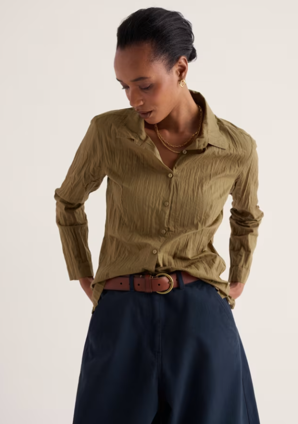 A woman wearing a textured olive green long-sleeve button-up shirt tucked into dark navy wide-leg trousers with a brown leather belt, photographed against a clean off-white studio background.