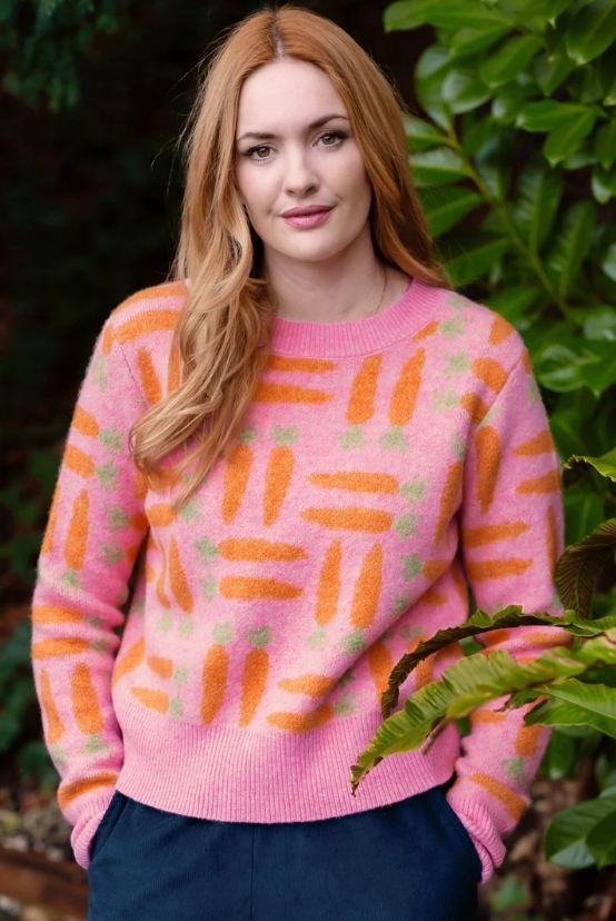 A woman with long wavy red hair wearing a pink knit sweater with an orange carrot pattern, standing outdoors in front of green foliage.