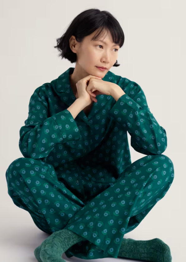 A woman sits cross-legged on a light grey studio floor wearing a dark green pyjama set with a small repeating circular pattern and matching green socks.
