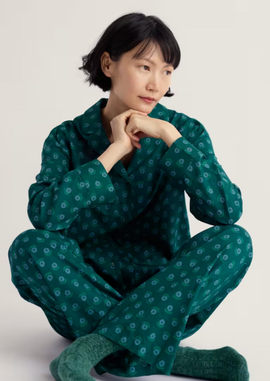 A woman sits cross-legged on a light grey studio floor wearing a dark green pyjama set with a small repeating circular pattern and matching green socks.