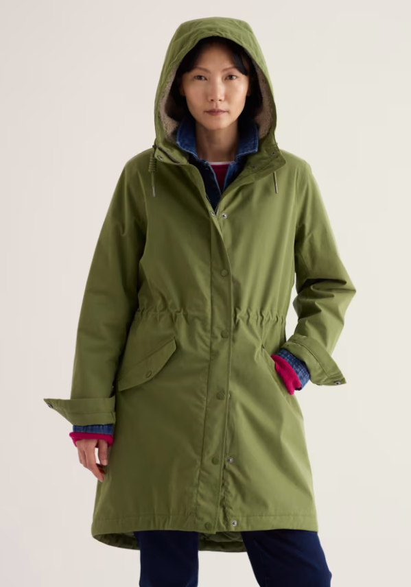 A woman wearing a hooded olive green coat with a drawstring waist and front pockets, layered over a blue plaid shirt and a red top, photographed against a plain light background.