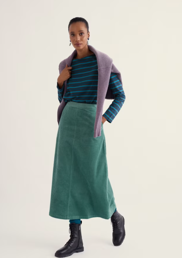 A model stands against a plain light beige studio background wearing a teal corduroy midi skirt, a dark blue and teal striped long-sleeve top, a lavender knit shrug draped over her shoulders, and black lace-up ankle boots.