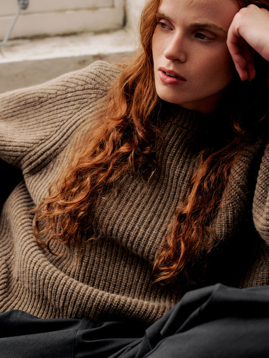A close-up lifestyle shot of a model with long, wavy red hair wearing a chunky, ribbed brown turtleneck sweater and dark gray pants, resting against a neutral concrete background.