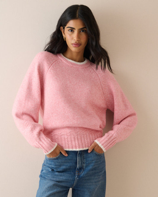 A woman wearing a soft pink knit crewneck sweater with white ribbed trim, paired with blue jeans, standing against a plain beige wall.