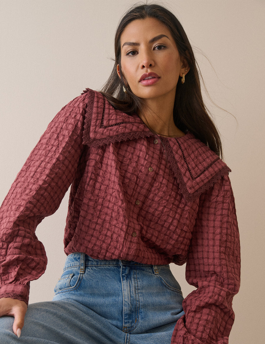 A woman wearing a long-sleeve crinkled blouse in a dusty rose and dark red checkered pattern with a large ruffled collar, paired with blue jeans, against a plain beige background.