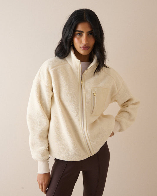 A woman modeling a cream-colored sherpa fleece zip-up jacket with gold-tone hardware, paired with dark brown pants, against a solid beige studio background.