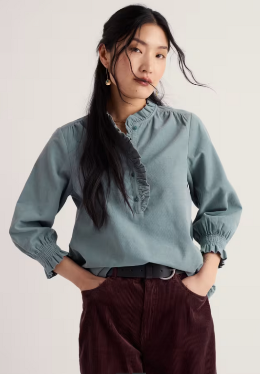A woman modeling a dusty blue long-sleeve blouse with a ruffled high collar and elasticized cuffs, paired with dark brown corduroy pants, against a plain off-white background.
