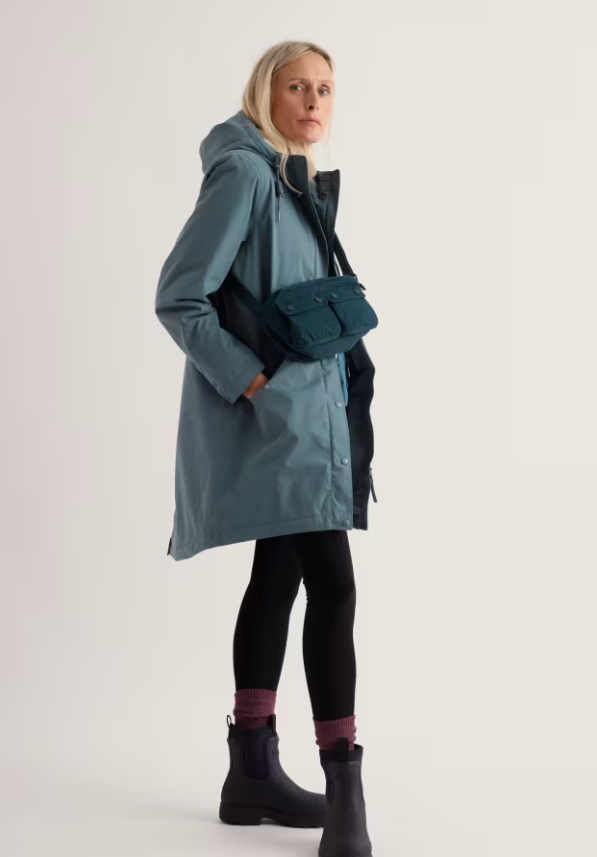 A woman modeling a slate blue hooded parka with black trim, wearing a matching blue crossbody bag, black leggings, and black boots against a plain light gray studio background.