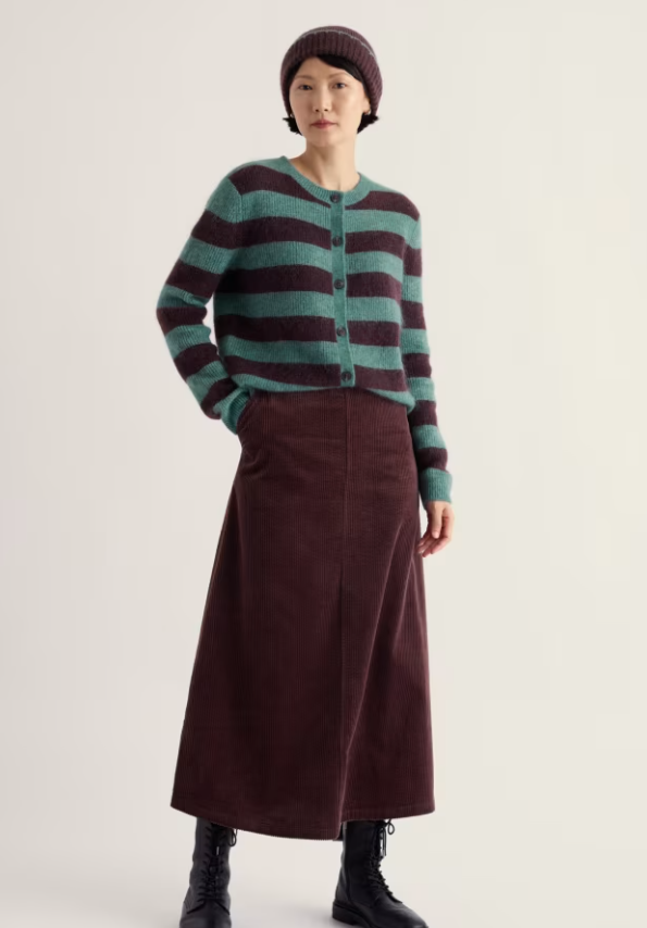 A model wearing a green and dark burgundy striped cardigan, a matching dark burgundy midi skirt, a dark burgundy beanie, and black ankle boots, standing against a plain light beige studio background.