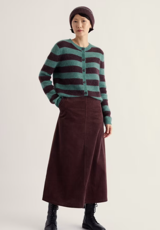A model wearing a green and dark burgundy striped cardigan, a matching dark burgundy midi skirt, a dark burgundy beanie, and black ankle boots, standing against a plain light beige studio background.
