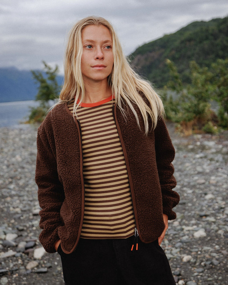A woman standing outdoors on a rocky lakeshore wearing a brown sherpa zip-front jacket over a beige and brown horizontally striped shirt, with dark pants and a cloudy mountain landscape in the background.